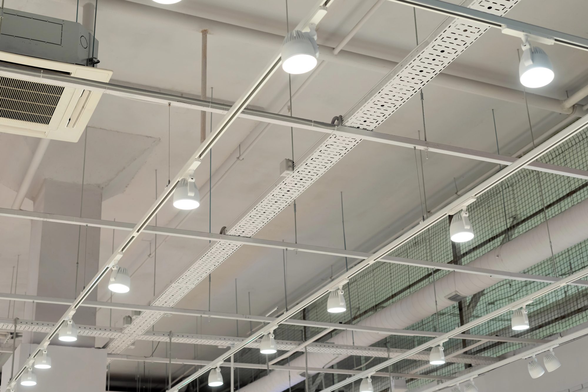 Ceiling with exposed pipes, air conditioning vent, cable trays, and multiple lit spotlights in a modern industrial-style interior.