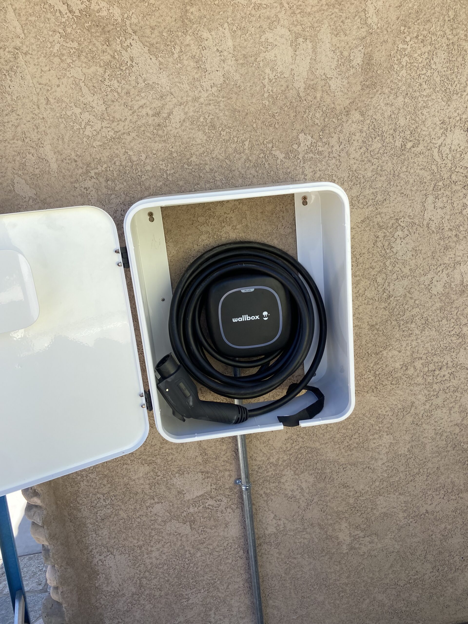 A Wallbox electric vehicle charger with its cable coiled is mounted inside a white box attached to a textured beige wall.