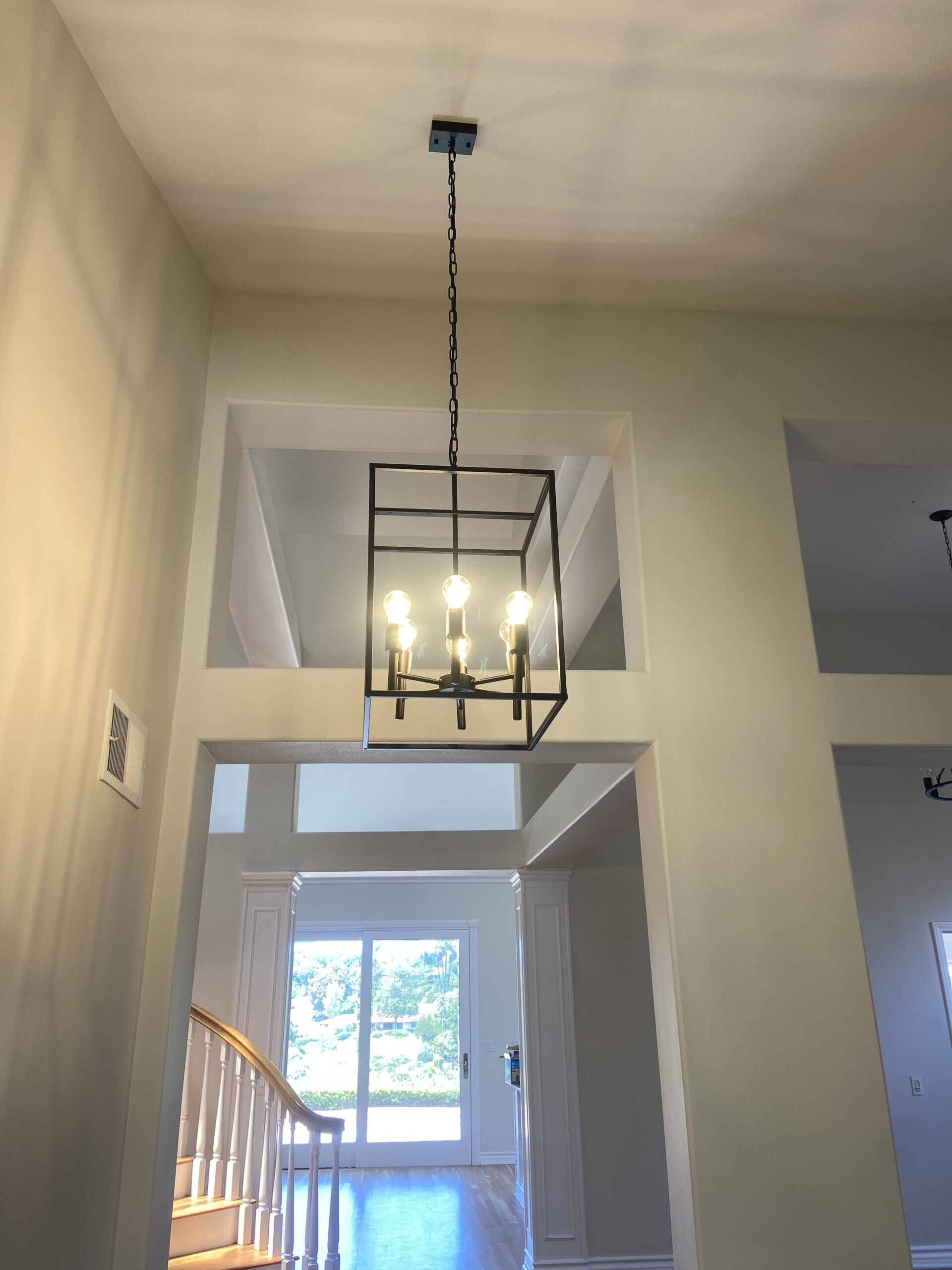 A black metal geometric pendant light with four exposed bulbs hangs from the ceiling in a bright entryway with light walls and natural sunlight coming through nearby windows.