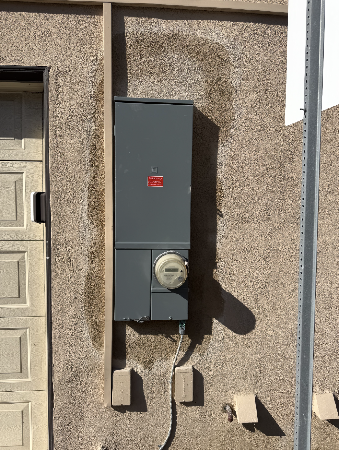 A gray electrical meter box is mounted on a stucco exterior wall next to a garage door, with conduit pipes running vertically and horizontally.