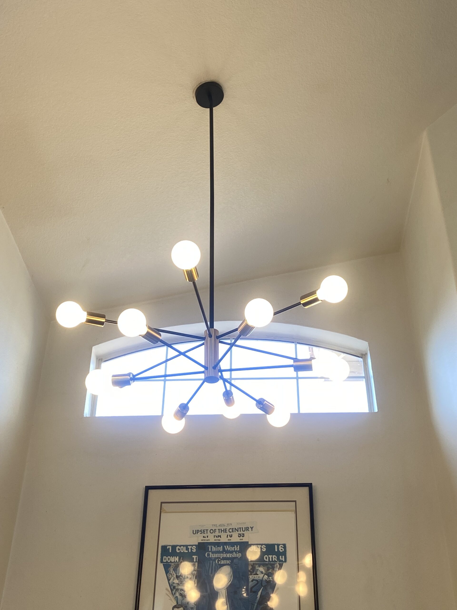 A modern sputnik chandelier with exposed bulbs hangs from the ceiling in a bright room with a small window and framed wall art below.