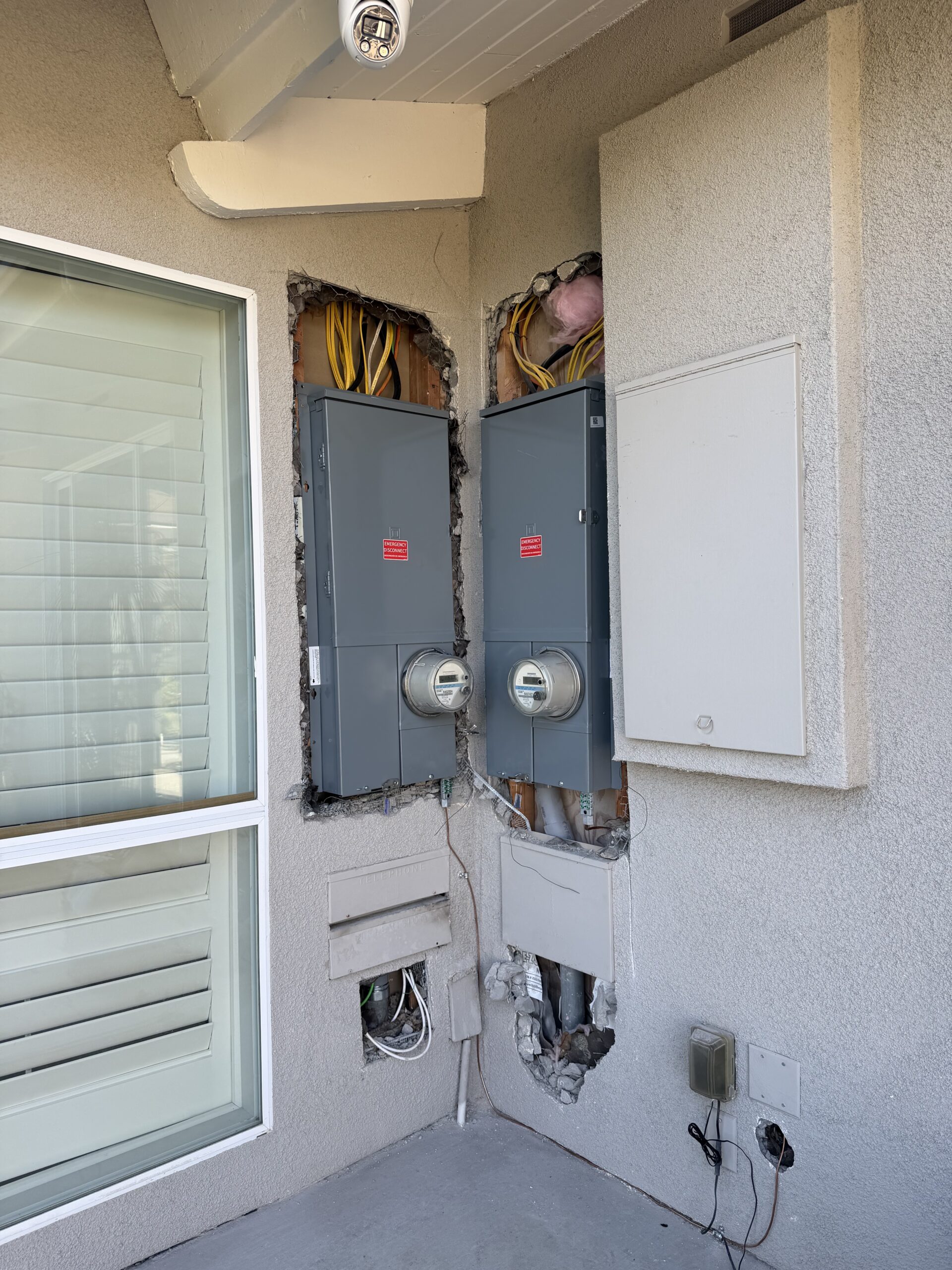 Two gray electrical meter panels installed in a wall with exposed wires and insulation; nearby is a window, a covered box, and some outlets.