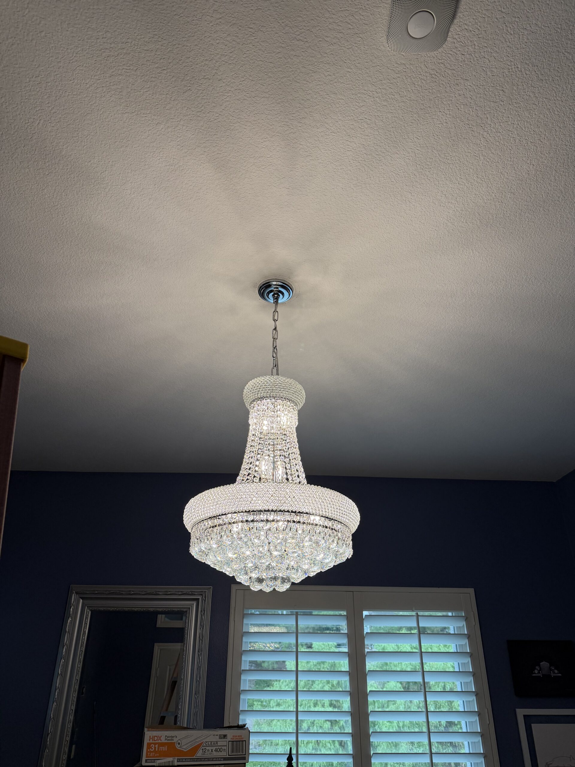 A crystal chandelier with multiple tiers hangs from the ceiling in a room with blue walls, a window with white shutters, and a large mirror.
